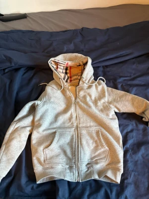Grå zip hoodie med Burberry-mönster - Snygg grå zip hoodie från Burberry med klassiskt rutigt foder i huvan och insidan. Hoodien har två stora fickor fram, dragsko i huvan och ribbade muddar. Perfekt för dig som vill ha en stilren men ändå lyxig look.
