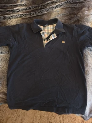 Marinblå pikétröja från Burberry - Klassisk marinblå pikétröja från Burberry med den ikoniska rutiga detaljen vid kragen och broderad logga på bröstet. Tröjan har korta ärmar och normal passform, perfekt för en stilren look. Tillverkad i mjuk bomull.