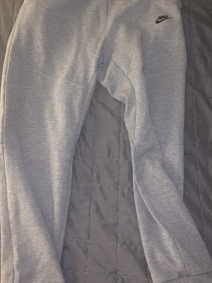 Grå mjukisbyxor från Nike - Snygga ljusgrå mjukisbyxor från Nike med klassisk logga på låret. Byxorna har en tapered pass sopassning och dragkedja på ena benet. Perfekta för chill eller träning, tillverkade i mjuk bomullsblandning för maximal komfort. Använt 3 gånger 