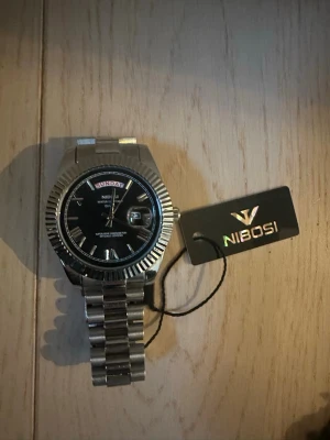 Silverfärgad klocka från Nibosi - Stilren klocka från Nibosi i silverfärgat rostfritt stål med svart urtavla. Klockan har dag- och datumvisning, markerade index och en klassisk länkarmband. Perfekt accessoar för dig som gillar en elegant och modern look.