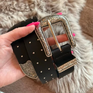Svart bälte med silverdetaljer - Snyggt svart bälte med bred rem i ”skinn” och dekorativa silverfärgade spännen och nitar. Bältet ger en cool western-vibe