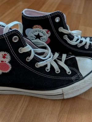 Svarta Converse All Star med patchar - Unika svarta Converse All Star high tops med vita snören och sula. Skorna har dekorativa blommiga och rutiga patchar runt loggan och på sidan, vilket ger dem en personlig och lekfull stil. Klassisk rund tå och kontrastsömmar. Perfekta för dig som vill sticka ut.