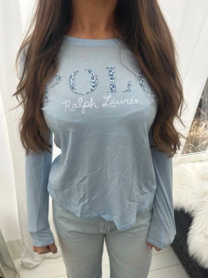 Ljusblå långärmad topp Polo Ralph Lauren - Ljusblå långärmad topp från Polo Ralph Lauren med rund halsringning. Framtill står det 'POLO Ralph Lauren' där bokstäverna har ett blommigt mönster i blå toner. Tunn och mjuk kvalitet, perfekt till jeans eller kjol. Barnstorlek men skulle säga att den passar till XS/S också🩵