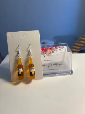 Örhängen med Corona-flaskor *gåva ingår* - Unika örhängen formade som små Corona-flaskor i gul plast med realistiska etiketter. Perfekt för dig som vill sticka ut och gillar roliga accessoarer. Krokar i silverfärgad metall. 