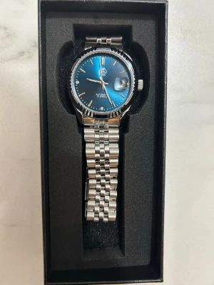 Klocka med blå urtavla - Snygg automatisk klocka med metallarmband i silver och en stilren blå urtavla. Klockan har datumvisning och klassisk rund form. Perfekt accessoar för dig som gillar en elegant och modern look.