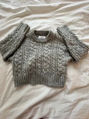 Grå kabelstickad cropped tröja - Säljer en grå kabelstickad cropped tröja med bred ribbad mudd i nederkant och vid ärmslut. Tröjan har rund halsringning och ett chunky mönster som ger en trendig vibe. Perfekt för lager på lager och riktigt snygg till höga jeans.🩶