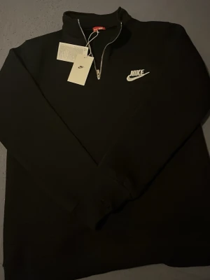Svart Nike tröja  - Svart långärmad tröja från Nike med vit broderad logga på bröstet. Tröjan har en praktisk half zip-dragkedja och hög krage. Tillverkad i mjukt material som passar perfekt till en avslappnad stil.