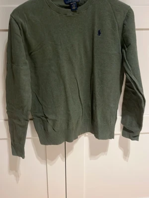 Olivgrön tröja från Ralph Lauren - Snygg olivgrön tröja från Polo Ralph Lauren i mjuk pima bomull. Tröjan har rund halsringning, långa ärmar och en liten broderad logga i mörkblått på bröstet. Perfekt för en stilren och avslappnad look.