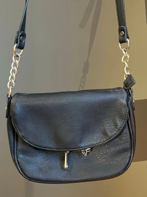 Svart axelväska med guldkedja - Stilren svart axelväska i skinnimitation med guldfärgade kedjedetaljer på axelremmen. Väskan har rundad form, lock och dragkedja framtill. Perfekt för dig som vill ha en enkel men trendig accessoar. Väl använd och vet inte vilket märke💛