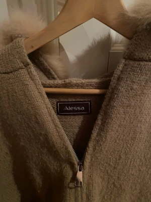 Beige/brun jacka från Alessa med fakepäls - Mysig jacka från Alessa med stor fluffig fuskpäls runt luvan och dragkedja framtill. Jackan har ribbade muddar. Använd ett fåtal gånger, lite nopprig, men sådant som man inte lägger märke till och som går bort med en noppermaskin!