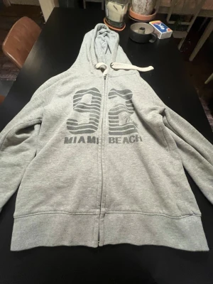Grå zip hoodie med Miami Beach-tryck - Säljer en grå zip hoodie med huva och vita dragsnören. På bröstet står det '92 MIAMI BEACH' i mörkgrått tryck. Hoodien har långa ärmar, ribbade muddar och är tillverkad i mjukt sweatshirtmaterial. Perfekt för chill dagar eller när du vill ha en avslappnad stil.