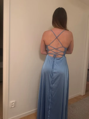Ljusblå långklänning med snörning - Snygg ljusblå långklänning i satinliknande material med smala axelband och öppen rygg med dekorativ snörning. Klänningen har en slits framtill som är ungefär upp till halva låret. Jag är 170cm lång och klänningen går ner till golvet. Perfekt för sommarens alla tillfällen.                            Priset är inte hugget i sten. 