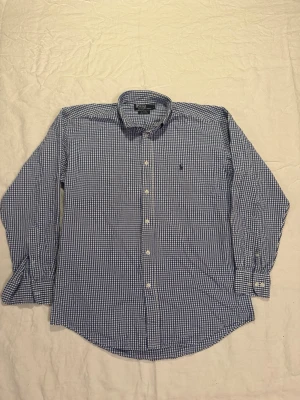 Blårutig skjorta från Polo Ralph Lauren - Klassisk blårutig skjorta från Polo Ralph Lauren med button-down krage och broderad logga på bröstet. Skjortan är tillverkad i 100% bomull och har långa ärmar samt vita knappar framtill. Perfekt för en clean och stilren look.