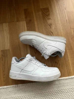 Nike Air Force 1 vita sneakers - Klassiska vita Nike Air Force 1 sneakers i skinn med låg profil och rund tå. Skorna har snörning, perforerade detaljer på tån och den ikoniska swoosh-loggan på sidan. Perfekta för en clean och stilren look. Finns ingen kartong till! DU VET VARFÖR DE ÄR BILLIGA