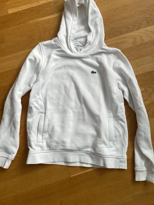 Vit hoodie från Lacoste Sport - Snygg vit hoodie från Lacoste Sport med klassisk krokodil-logga på bröstet. Hoodien har en mjuk bomullskänsla, fickor på sidorna och ribbade muddar. Perfekt för en clean och sportig look.