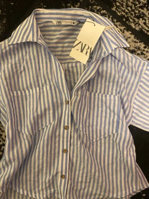 Blårandig skjorta från Zara - En snygg blårandig skjorta från Zara med korta ärmar och fickor. Skjortan har krage och knappar framtill. HELT OANVÄND MED PRISLAPP KVAR.