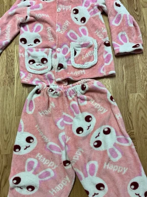Rosa pyjamas med kaninmotiv - Supermjuk och fluffig rosa pyjamas med söta vita kaniner och texten 'Happy' över hela. Setet består av långärmad tröja med knappar och två fickor samt matchande byxor med resår i midjan. Perfekt för mysiga hemmakvällar och chill dagar.byxorna är 150 cm långa 