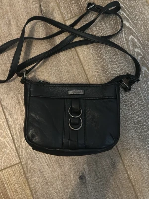 Svart axelväska  - Snygg svart axelväska i skinn med justerbar axelrem. Väskan har silverfärgade metalldetaljer framtill och en praktisk dragkedja på baksidan. Perfekt storlek för det viktigaste och enkel att matcha till olika outfits.