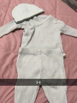 Ribbat set med mössa för baby - Mysigt ribbat loungewear-set för baby i ljusgrått, består av långärmad body med tryckknappar, matchande byxor och en söt mössa. Perfekt för chill dagar hemma eller som mjuk pyjamas. Materialet känns mjukt och stretchigt mot huden.