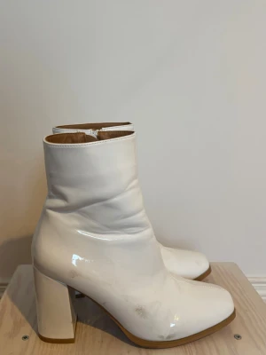 Vita stövletter med klack - Snygga vita stövletter i glansigt material med fyrkantig tå och bred hög klack. Dragkedja på insidan för enkel påtagning. Perfekta för dig som vill sticka ut med ett par trendiga boots.