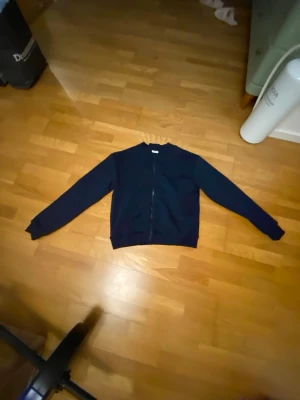 Mörkblå bomberjacka från Kappahl - Snygg mörkblå bomberjacka från Kappahl med klassisk rak passform och dragkedja framtill. Jackan har ribbade muddar vid ärmslut och nederkant, vilket ger en clean och stilren look. Perfekt för dig som vill ha en enkel och tidlös jacka.