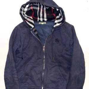 Marinblå hoodie från Burberry - Snygg marinblå hoodie från Burberry med dragkedja och klassiskt rutigt foder i huvan. Hoodien har två fickor framtill, justerbar dragsko i huvan och diskret Burberry-logga broderad på bröstet. Perfekt för en stilren och avslappnad look.