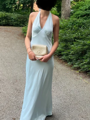 Ljusblå långklänning med öppen rygg - Snygg ljusblå långklänning i satin med halterneck och v-ringning. Klänningen har en figurnära passform på överkroppen och går ut nätt vid benen. Knyte i både nacke och rygg vilket ger den en vacker och modern look. Perfekt till bal, fest eller sommar tillfällen.                            Använd en gång! 