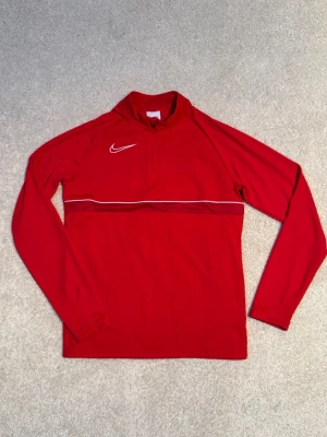 Röd Nike träningströja med dragkedja - Snygg röd långärmad träningströja från Nike med vit logga på bröstet och en tunn vit linje över bröstet. Tröjan har halv dragkedja framtill och är tillverkad i ett lätt och stretchigt material som passar perfekt för träning och till vardags. Juniorstorlek L (147-158cm)