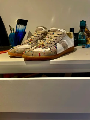 Maison Margiela sneakers med färgstänk - Unika sneakers från Maison Margiela med beige och vita paneler, klassisk gummisula och färgstänk i rött, blått, gult och grönt. Skorna har snörning och en rund tå. Perfekta för dig som vill sticka ut med din stil. Finns box med. Pris ej hugget i sten