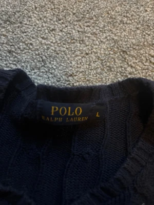 Mörkblå kabelstickad tröja Polo Ralph Lauren - Mörkblå kabelstickad tröja från Polo Ralph Lauren med rund halsringning och klassisk orange logga broderad på bröstet. Tröjan har långa ärmar och ribbade muddar. Perfekt för en stilren och tidlös look.