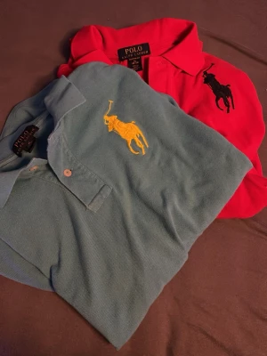 Pikétröjor från Polo Ralph Lauren - Två klassiska pikétröjor från Polo Ralph Lauren, en i ljusblått med gul broderad logga och en i rött med svart broderad logga. Båda har krage, knappar framtill och är perfekta för en stilren look.