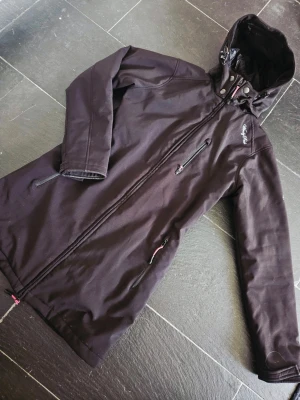 Svart vindjacka från Windy Peak - Snygg svart vindjacka från Windy Peak med huva och dragkedja framtill. Jackan har två sidofickor med dragkedja och en bröstficka. Materialet är slitstarkt och jackan har en längre passform som skyddar mot vind. Perfekt för dig som vill ha en stilren och funktionell jacka.