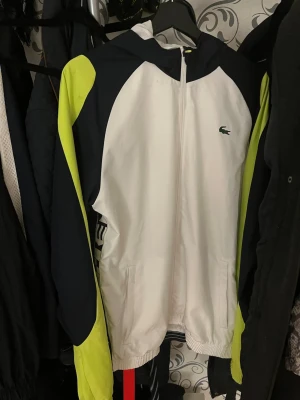 Lacoste vindjacka med huva - Snygg vindjacka från Lacoste i vitt, mörkblått och neongult. Jackan har huva, dragkedja framtill och två fickor. Ikoniska Lacoste-loggan på bröstet. Perfekt för dig som vill sticka ut med sportig stil.