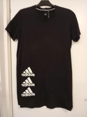 Svart Adidas t-shirt klänning med logga - Svart t-shirt klänning från Adidas med tre vita Adidas-loggor tryckta nertill på framsidan. Klassisk rund halsringning och korta ärmar. Tillverkad i mjuk bomull som känns skön mot huden. Perfekt för en sportig och avslappnad stil.