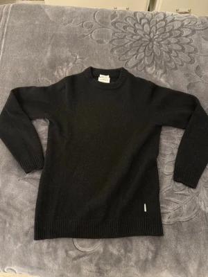 Svart stickad tröja från Lindbergh - Svart stickad tröja från Lindbergh i relaxed fit, tillverkad i mjuk lammull. Tröjan har rund halsringning, långa ärmar och ribbade muddar vid ärmslut och nederkant. Perfekt för kalla dagar och enkel att matcha med olika outfits.