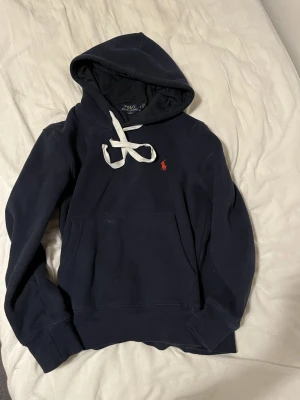 Marinblå hoodie från Polo Ralph Lauren - Mörkblå hoodie från Polo Ralph Lauren i slim fit-modell. Klassisk huva med vita dragsnören och känguruficka framtill. Ikoniska röda logotypen broderad på bröstet. Tillverkad i mjuk bomullsblandning för skön känsla.