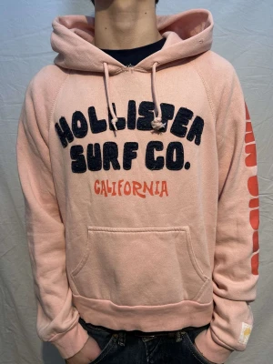 En fet rosa hollister hoodie!! - En snygg och unik rosa vintage hoodie med najs tryck!!!  Märke:Hollister!! OBS UNISEX så passar perfekt till både killar och tjejer. Storlek S. Modellen är 163cm lång och väger 53kg. Skick 7/10 pågrund av en väldigt liten defekt som syns på bild 5 obs syns knappt