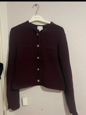 Vinröd kofta med guldknappar - Mysig vinröd kofta från Mac Scott med snygga guldknappar framtill. Koftan har en rak, boxig passform, rund halsringning och två dekorativa bröstfickor. Stickad struktur som ger en trendig look och passar perfekt till lager på lager.