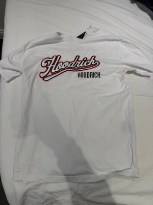 Vit Hoodrich t-shirt med tryck - Säljer en vit t-shirt från Hoodrich med stort tryck fram och bak. Framtill står det 'Hoodrich' i röd och svart text, och baktill finns ett stort 'Hoodrich Hood Series'-tryck. Klassisk passform och rund hals. Perfekt för dig som gillar streetwear.
