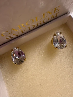 Caroline Svedbom droppformade örhängen - Snygga örhängen från Caroline Svedbom med droppformade, klara kristaller infattade i guldfärgade detaljer. Perfekta för att ge en glittrig touch till din look. Kommer i originalask.