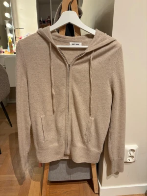 Beige hoodie i kashmir från Soft Goat - Mysig beige hoodie i mjuk kashmir från Soft Goat. Dragkedja framtill, två fickor och justerbar huva med snören. Perfekt för en avslappnad och stilren look. Långa ärmar och ribbade muddar ger en skön passform.