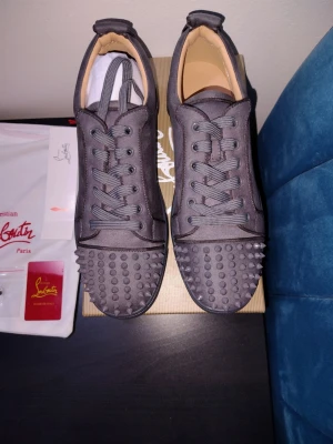 Svarta Christian Louboutin sneakers - Snygga svarta sneakers från Christian Louboutin med ikoniska röda sulor och coola nitar på tån. Skorna har snörning och är tillverkade i textil och skinn för en lyxig känsla. Perfekta för dig som vill sticka ut med din stil.