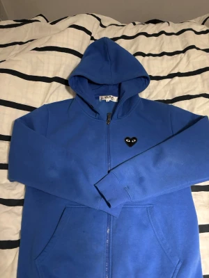 Blå Comme des Garçons PLAY hoodie - Blå hoodie med dragkedja från Comme des Garçons PLAY. Har den ikoniska svarta hjärtloggan med ögon på bröstet och en stor version på ryggen. Utrustad med huva och fickor framtill. Perfekt för dig som gillar streetwear och vill sticka ut.