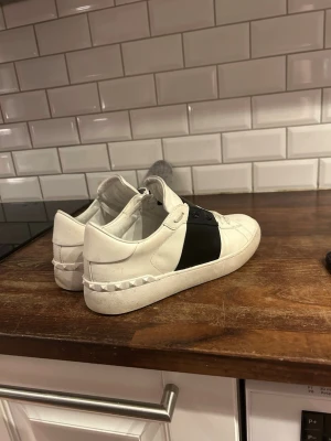 Valentino Garavani vita sneakers - Snygga vita sneakers från Valentino Garavani med svart band över vristen och ikoniska nitar på sulan. Skorna har snörning och är tillverkade i skinn för en lyxig känsla. Perfekta för dig som vill ha en stilren och exklusiv look. Box medföljer
