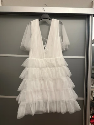 Vit volangklänning med snörning - Supersöt vit kortklänning med flera lager volanger och transparenta kortärmar. Djup v-ringning både fram och bak med snörningsdetalj i ryggen. Tillverkad i ett tunt, skirt material som ger en fluffig och luftig känsla.