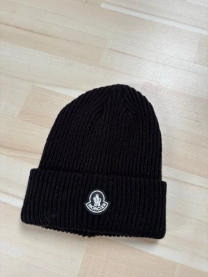 Svart ribbstickad mössa - Snygg svart ribbstickad mössa från med uppvikt kant och klassisk logotyp framtill. Tillverkad i mjukt material som håller värmen och ger en clean look. Perfekt accessoar för kalla dagar och streetwear-stil.