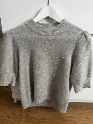 Grå stickad tröja med pärlor - Snygg grå stickad tröja med korta puffärmar och dekorativa pärlor framtill. Tröjan har ribbad halsringning, ärmslut och nederkant. Perfekt för dig som vill ha en trendig och mysig look med det lilla extra.