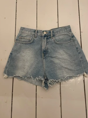 Blåa högmidjade jeansshorts - Säljer ett par ljusblå högmidjade jeansshorts med råa, fransiga kanter. Klassisk femficksmodell med knapp och dragkedja framtill. Perfekta för sommaren och har en loose passform för en avslappnad stil.