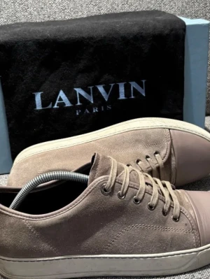 Beige sneakers från Lanvin i mocka - Snygga beige sneakers från Lanvin med ovandel i mjuk mocka och tåparti i matchande beige skinn. Klassisk rund tå, snörning och vit platt sula ger en clean och stilren look. Perfekta för dig som gillar exklusiva och tidlösa sneakers.