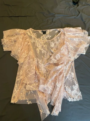 Ljusrosa spetsblus med volang - Romantisk ljusrosa blus i transparent spets med blommigt mönster. Blusen har volangdetaljer och omlott-design som knyts i sidan. Perfekt för dig som vill ha en feminin och trendig topp med korta ärmar.